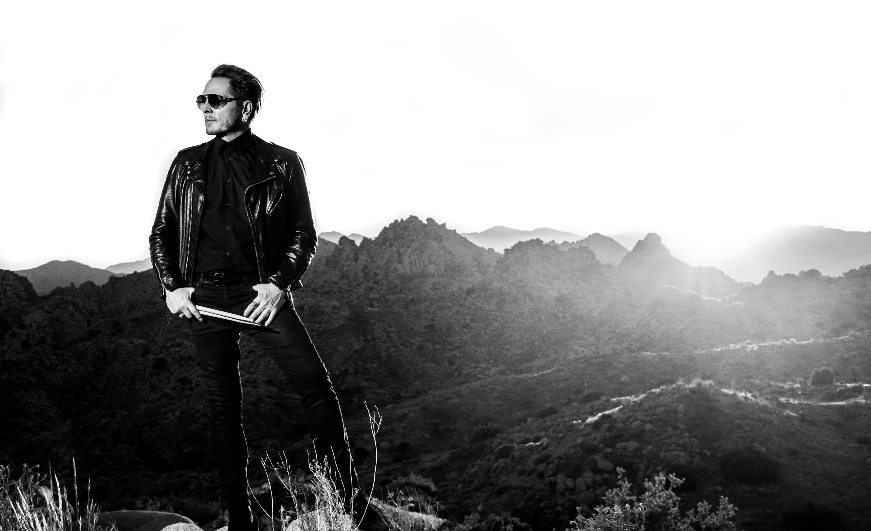 Matt Sorum black and white portrait on hill top in desert valley wearing sunglasses while holding drumsticks thumbs in his denim pockets