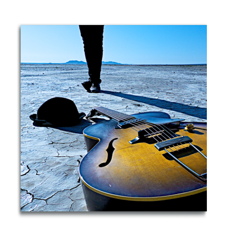 Fine art square color photographic print on metal Gibson acoustic vintage guitar lying on desert dried lakebed in foreground with hat beside it legs with feet crossed in background