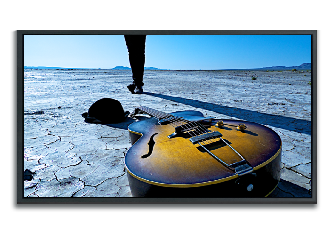 Fine Art color photography Gibson acoustic guitar lying in foreground on cracked dried desert lakebed hat beside it legs standing crossed behind it against blue sky