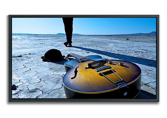 Fine Art color photography Gibson acoustic guitar lying in foreground on cracked dried desert lakebed hat beside it legs standing crossed behind it against blue sky