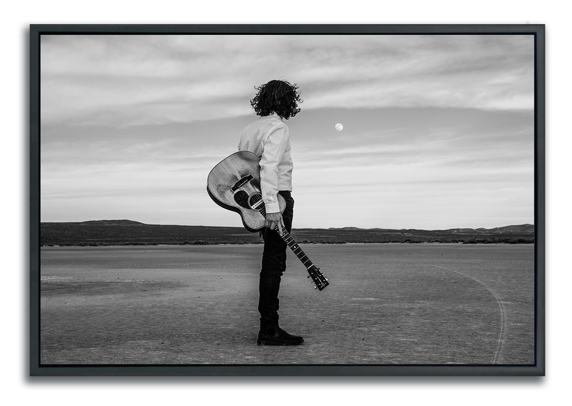 Black and white Fine Art Photography man standing in desert holding Gibson acoustic guitar back to camera staring at moon above the horizon