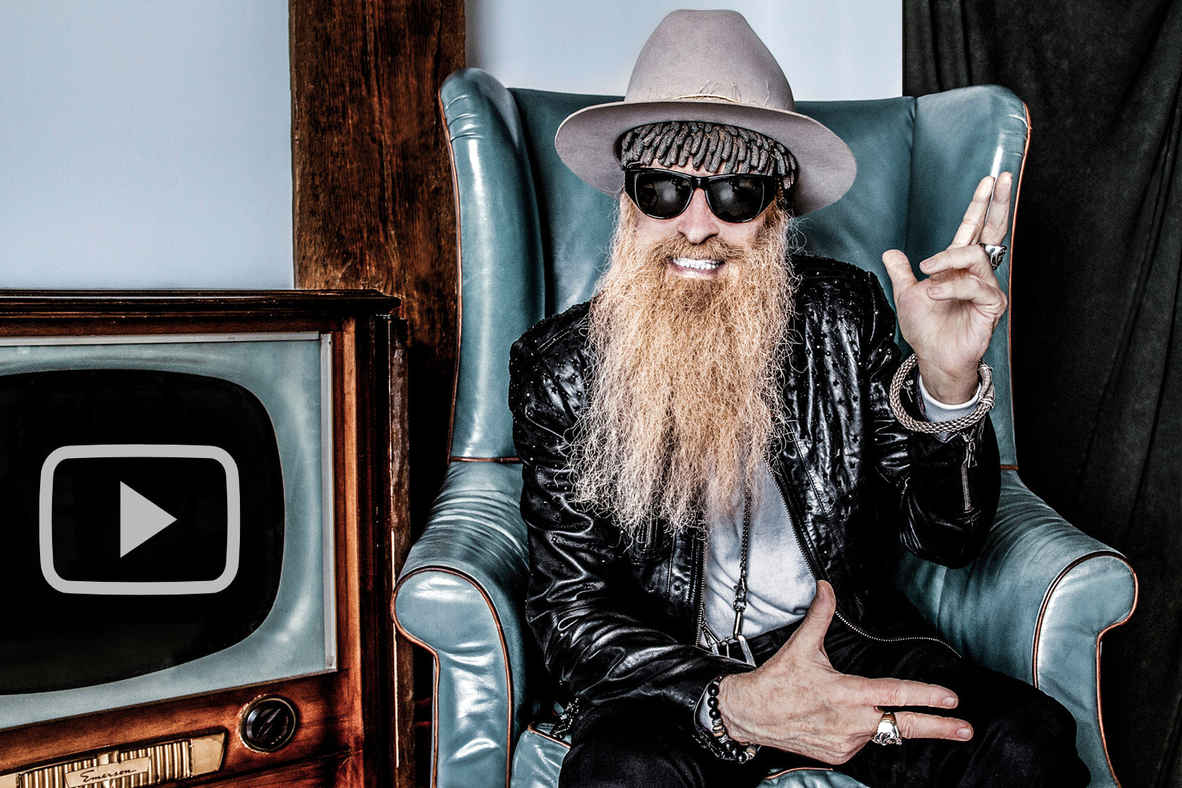 Billy F Gibbons portrait sitting in blue chair beside old television smiling and pointing two fingers on each hand one sideways and one up in the air