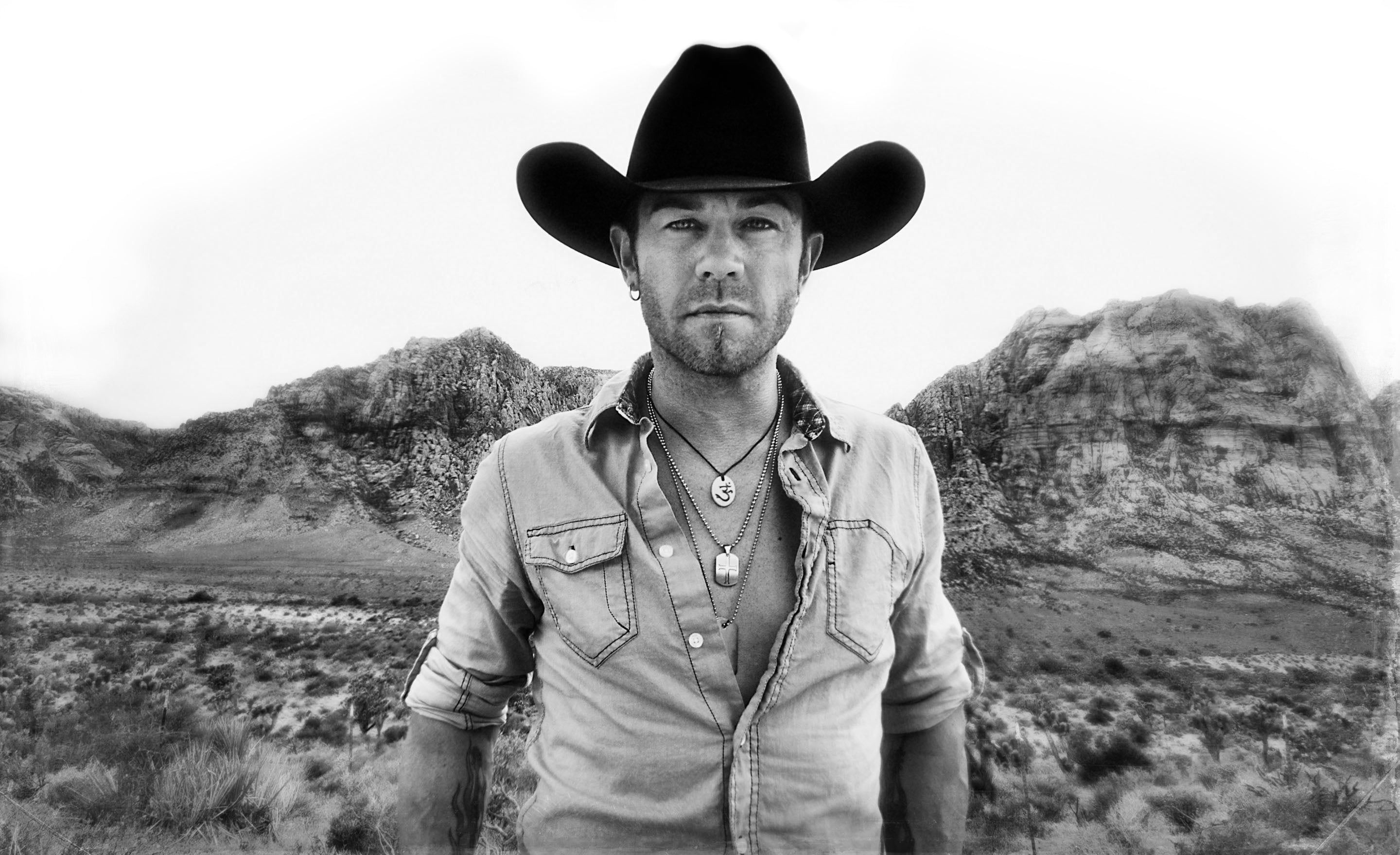 Black and white portrait country music artist Aaron Pritchett standing in front of desert canyon wearing black cowboy hat and denim shirt