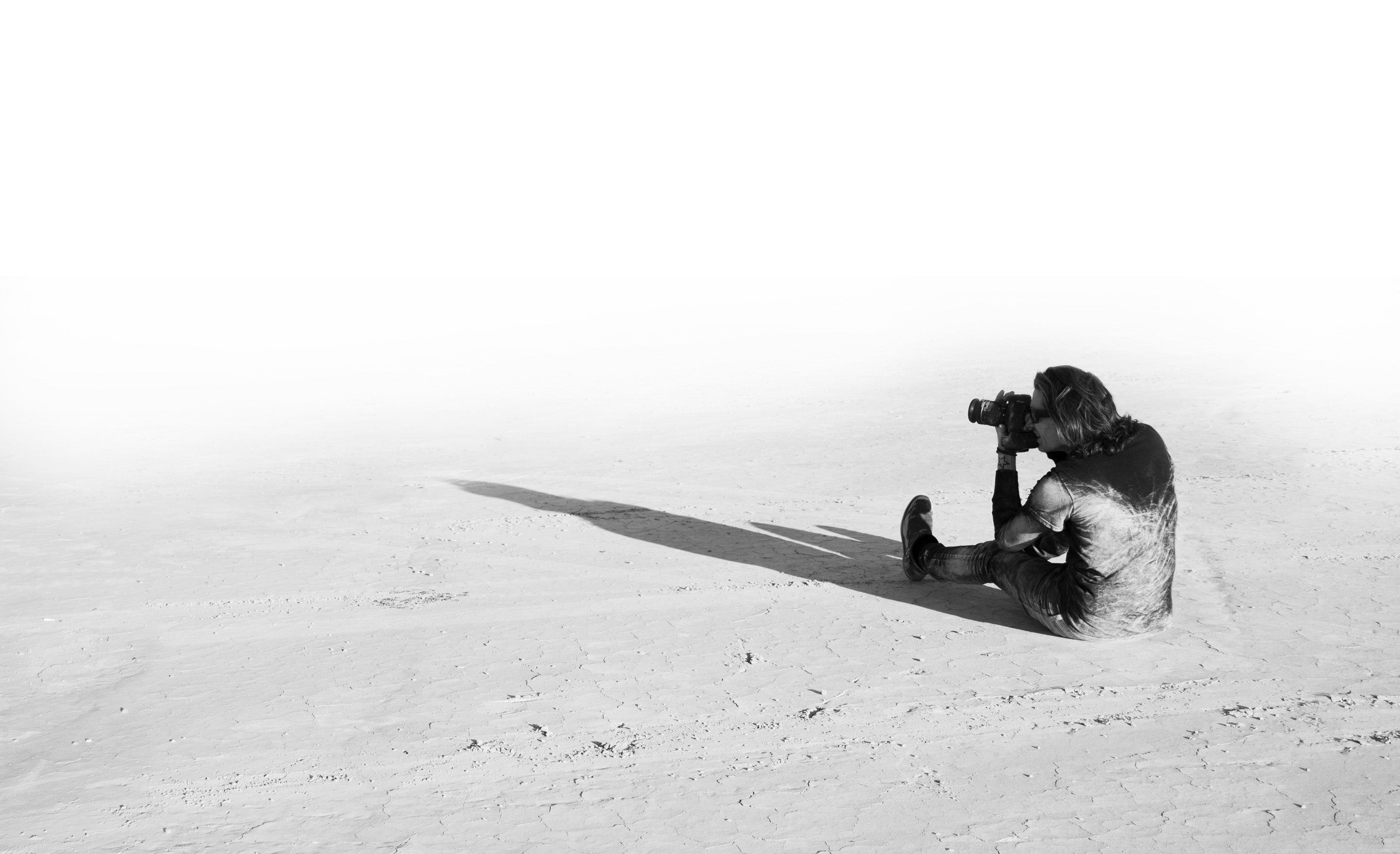 Fine Art Photographer Mark Maryanovich sitting on desert floor sand on his back looking through back of camera in black and white