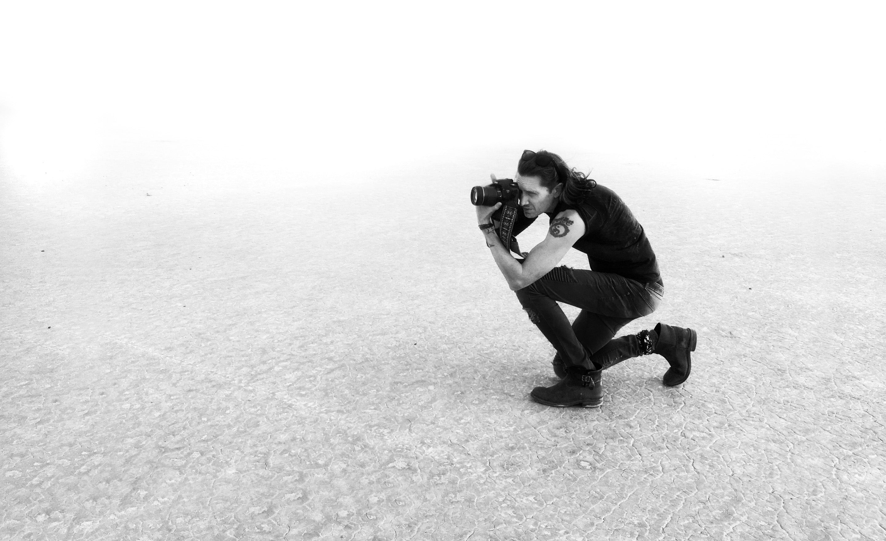 Fine art photographer Mark Maryanovich kneeling on dried lake bed floor looking through back of camera in black and white
