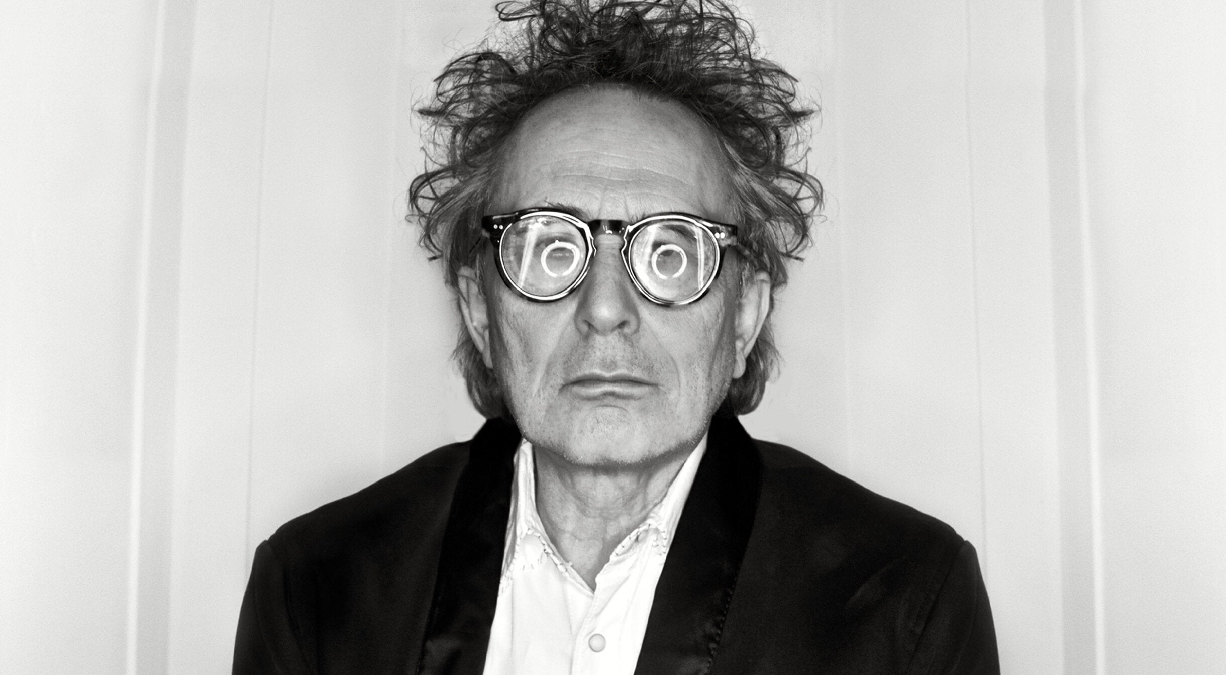 Black and white portrait musician Marc Jordan with swirl of circle light in reflection of his glasses while standing against white backdrop