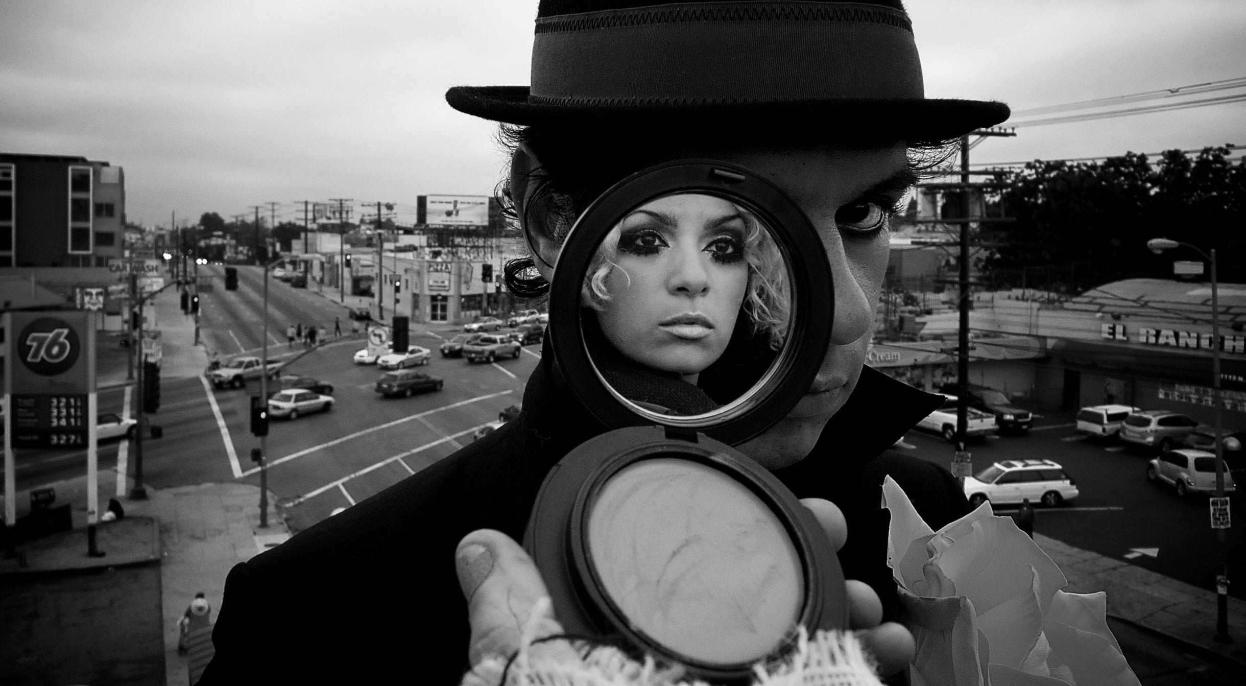 Black and white portrait music duo male band member standing on rooftop wearing hat makeup compact covering one side of his face image of female band member in reflection of mirror busy street in background
