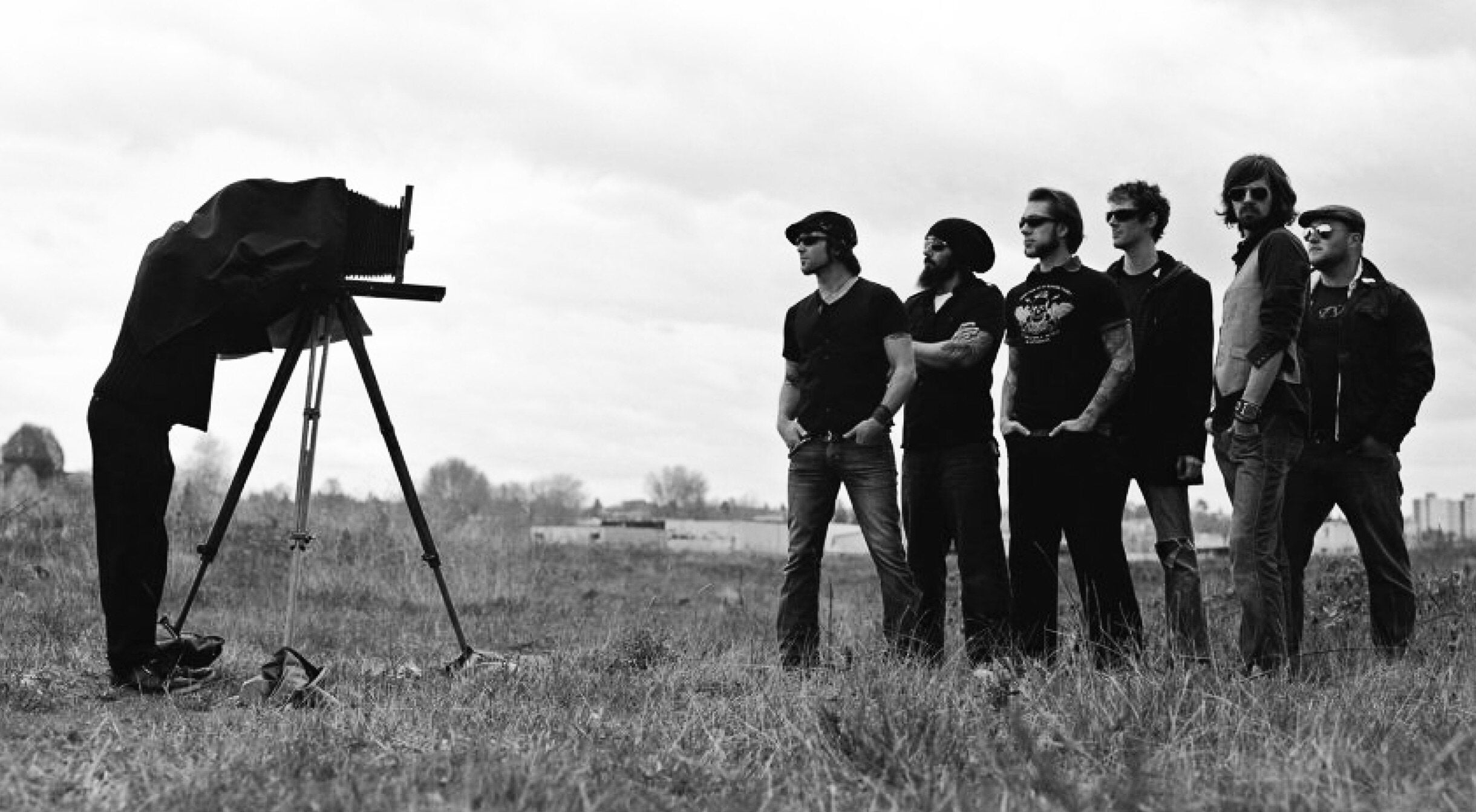 Black and white image seven members of The Matinee band standing in field facing old fashioned camera photographer under cloth taking photo