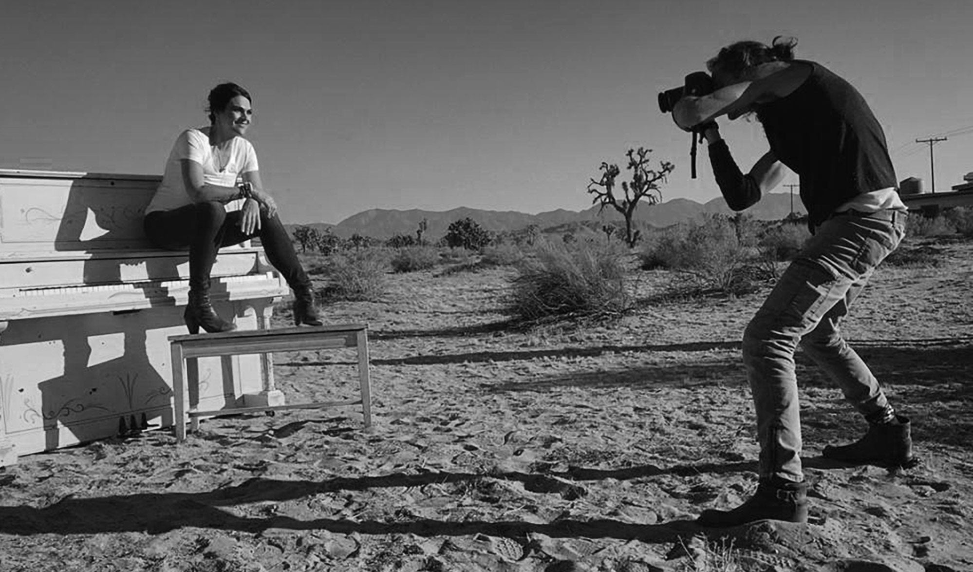 Fine Art Portrait Photography behind the scenes black and white photo Mark Maryanovich photographing Leslie Cours Mather sitting on piano placed in middle of California desert
