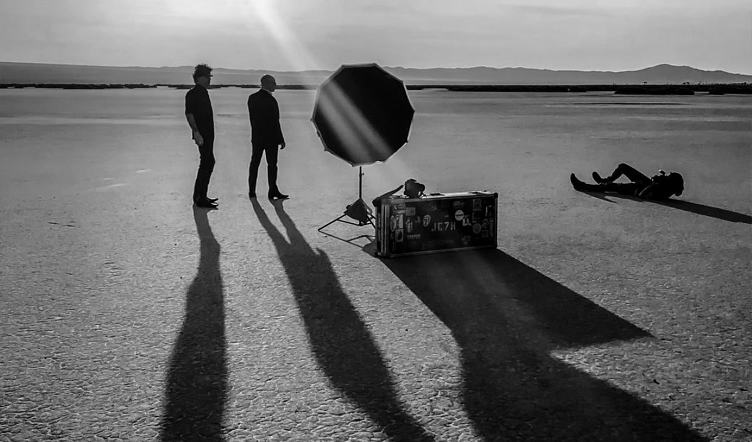 Fine art portrait photographer Mark Maryanovich black and white behind the scenes with duo 7Horse in El Mirage light set up two band members posing while Mark lays on desert floor photographing them with sun streak and long shadows