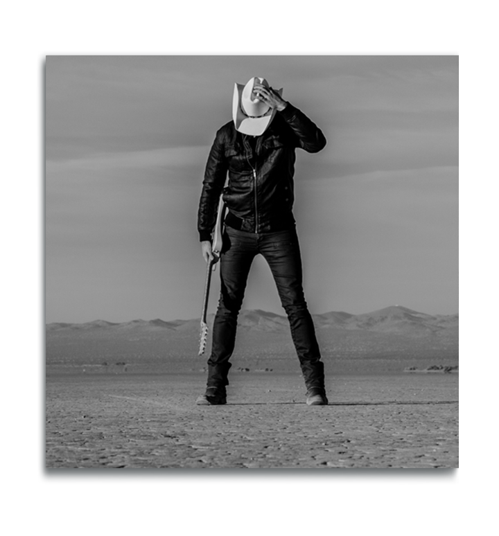 Black and white square metal print man standing in desert holding guitar head down holding onto his white cowboy hat covering his face