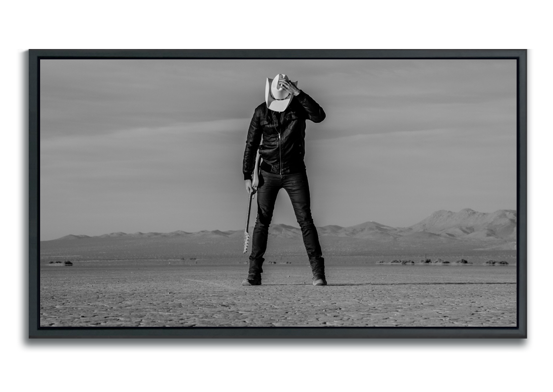 Black and white fine art photographic metal print man standing in desert holding guitar head down with hand keeping his white cowboy hat in place