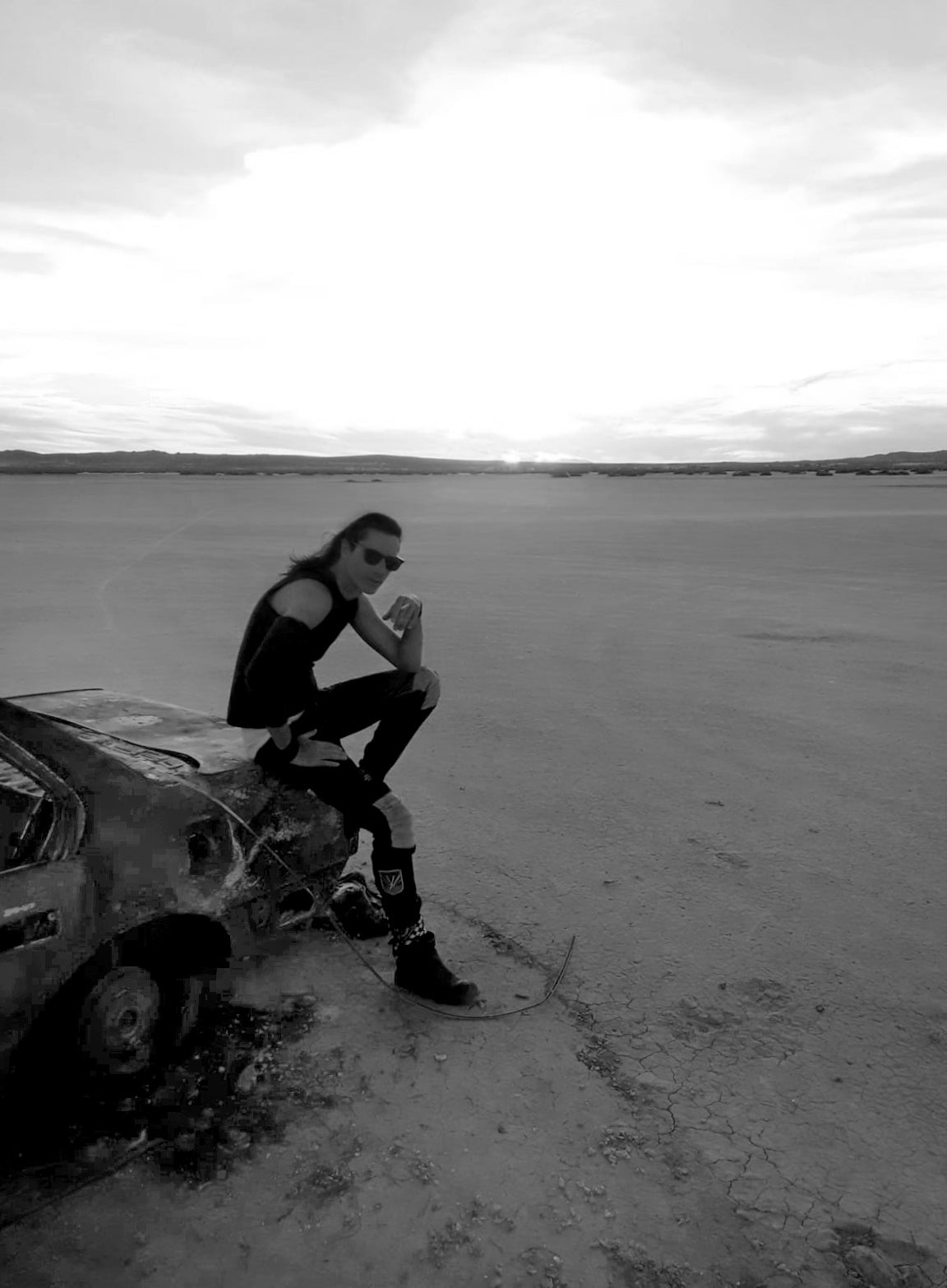 Fine art photographer Mark Maryanovich wearing sunglasses sitting on trunk of burned out car parked in middle of desert in black and white