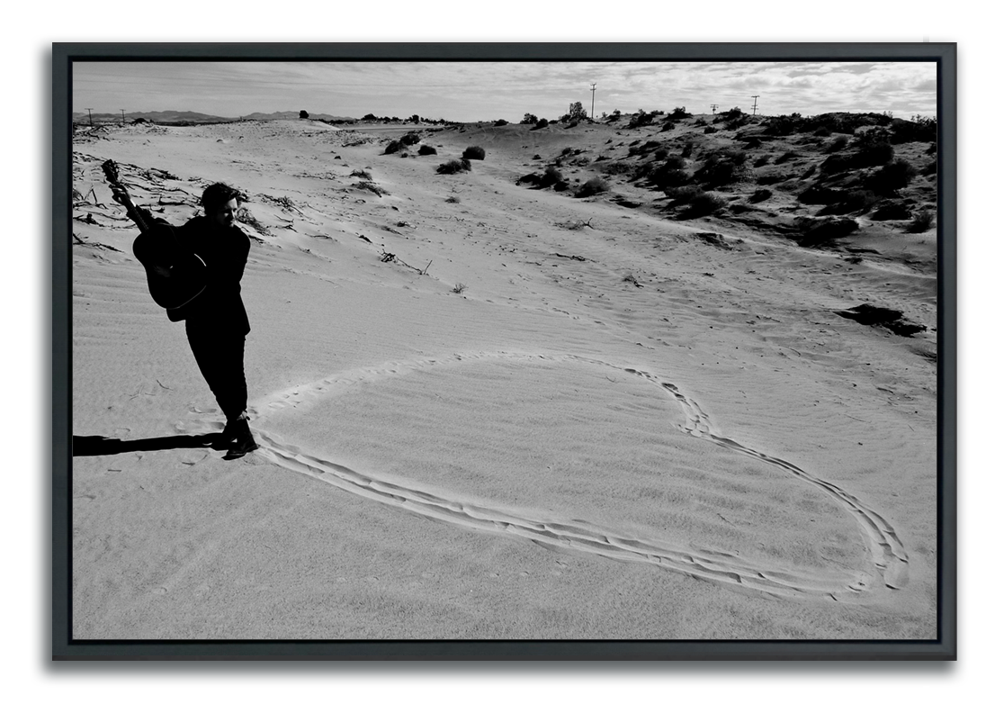 Black and white fine art photography men standing with guitar at the pint of a heart drawn in sand