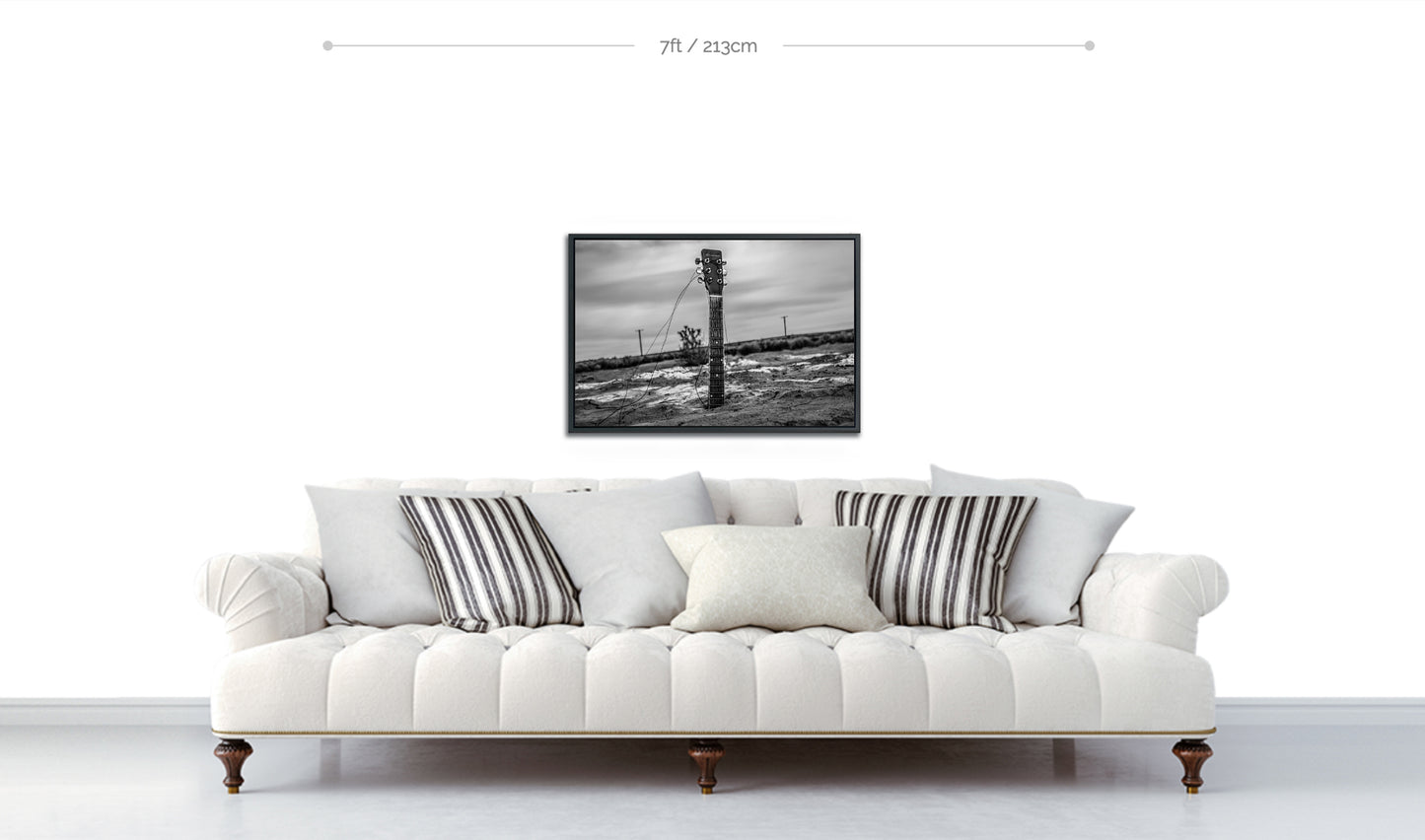 Black and white guitar neck protruding from body buried in desert sand framed photographic print displayed above white sofa 