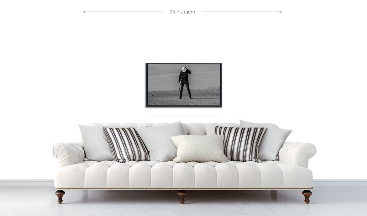 Black and white fine art framed metallic print hanging on white wall above sofa featuring man standing in desert holding guitar head down with hand keeping his white cowboy hat in place